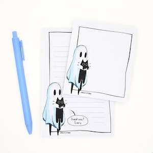 May include: A light blue retractable pen next to two white paper notepads featuring a cartoon ghost holding a black cat. The bottom notepad includes horizontal lines and a speech bubble with the text "Deadlines? Scary" and "Caroljy Studios".