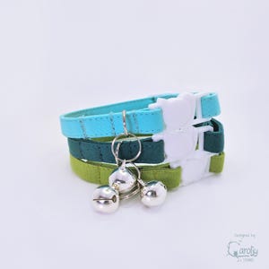May include: A stack of three fabric collars in shades of light blue, dark teal, and lime green. Each collar features a white plastic buckle shaped like a cat head and a silver metal ring holding two small, round, shiny silver jingle bells.