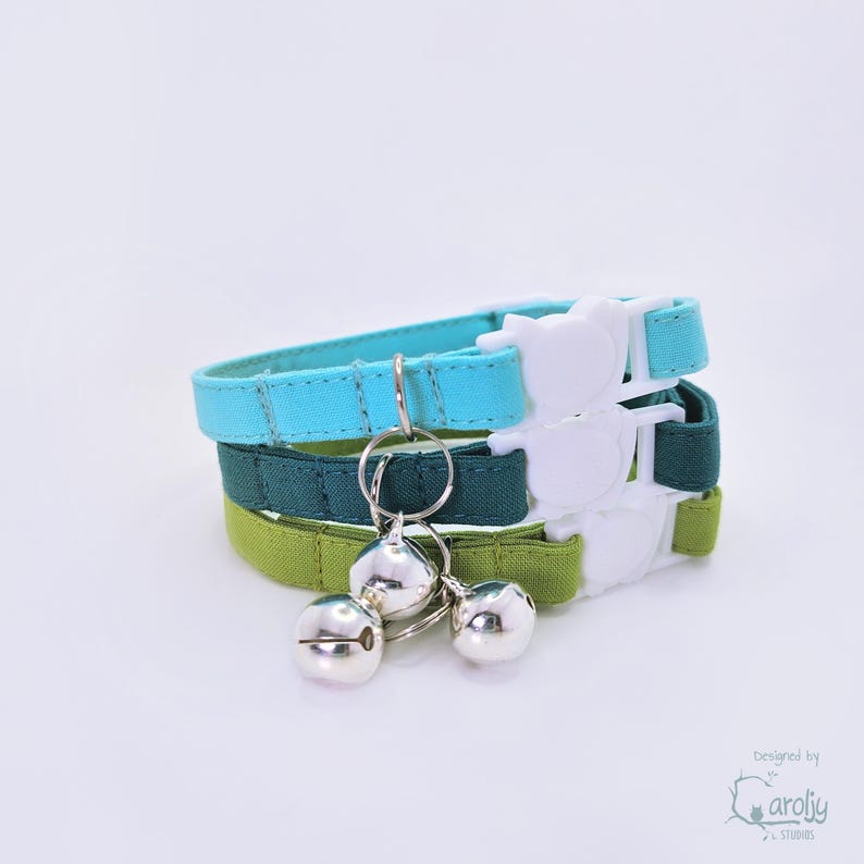 May include: A stack of three fabric collars in shades of light blue, dark teal, and lime green. Each collar features a white plastic buckle shaped like a cat head and a silver metal ring holding two small, round, shiny silver jingle bells.