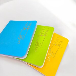 May include: Three notebooks in blue, green, and yellow. Each notebook features a gold illustration of a cat. The notebooks are stacked, with the blue notebook on top, followed by the green and yellow notebooks.