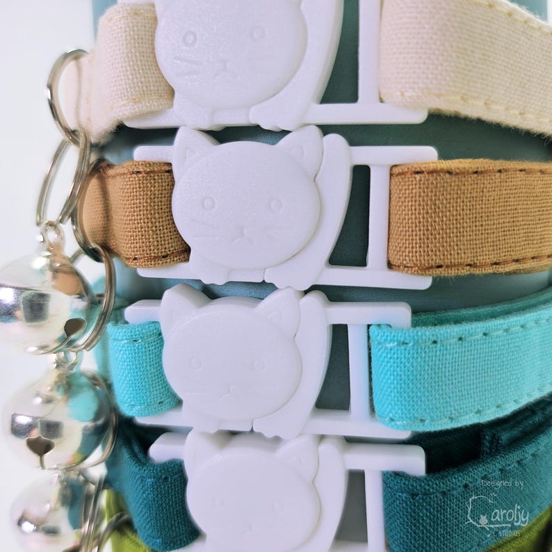 May include: A stack of four fabric collars in cream, tan, turquoise, and dark green, each featuring a white plastic buckle shaped like a cat face. Each collar includes a silver metal split ring attached to a small, round silver jingle bell.