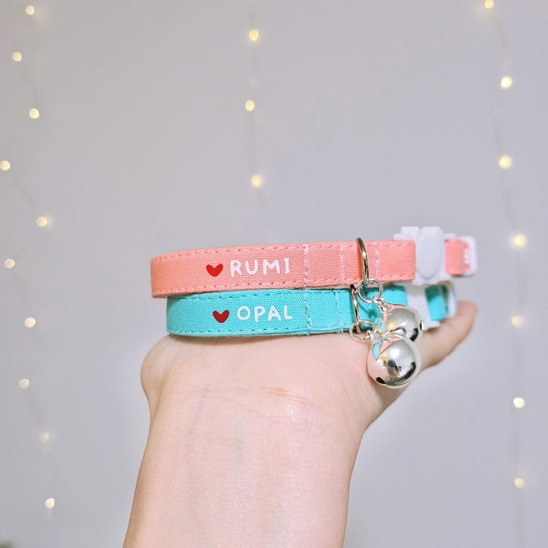 May include: Two fabric pet collars, one coral pink and one turquoise blue, each featuring a white plastic buckle, a silver metal bell, and a small red heart icon next to the white printed names RUMI and OPAL respectively.
