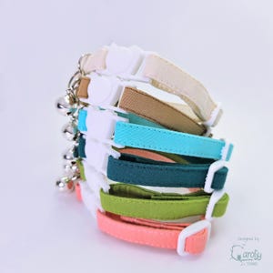 May include: A stack of six fabric pet collars in cream, tan, turquoise, dark teal, lime green, and coral. Each collar features a white plastic side-release buckle, an adjustable slider, and a small silver-toned metal bell attached to a metal ring.