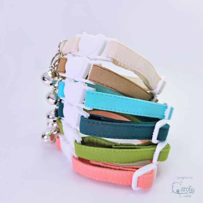 May include: A stack of six fabric pet collars in cream, tan, turquoise, dark teal, lime green, and coral. Each collar features a white plastic side-release buckle, an adjustable slider, and a small silver-toned metal bell attached to a metal ring.