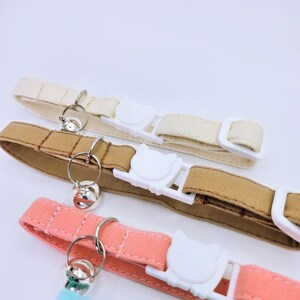 May include: Three fabric pet collars in cream, tan, and coral pink. Each collar features a white plastic buckle shaped like a cat head, a silver metal split ring, and a small silver bell. The collars include a white plastic slider for adjustment.