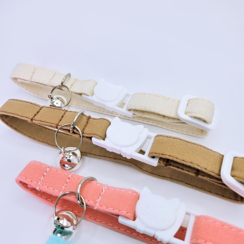 May include: Three fabric pet collars in cream, tan, and coral pink. Each collar features a white plastic buckle shaped like a cat head, a silver metal split ring, and a small silver bell. The collars include a white plastic slider for adjustment.