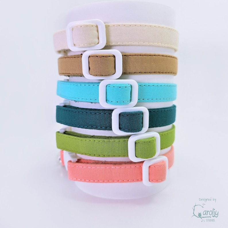 May include: A stack of six fabric collars in cream, tan, turquoise, dark teal, lime green, and coral. Each collar features a white plastic buckle and a matching white plastic slider. The text "Designed by caroljy STUDIOS" appears in the bottom right.