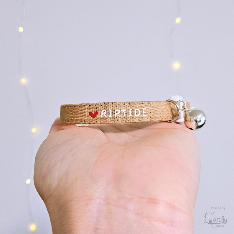 May include: A tan fabric pet collar featuring a small red heart icon followed by the white text RIPTIDE. The collar includes a silver-toned metal bell attached to a metal ring, with a white plastic buckle closure visible on the side.