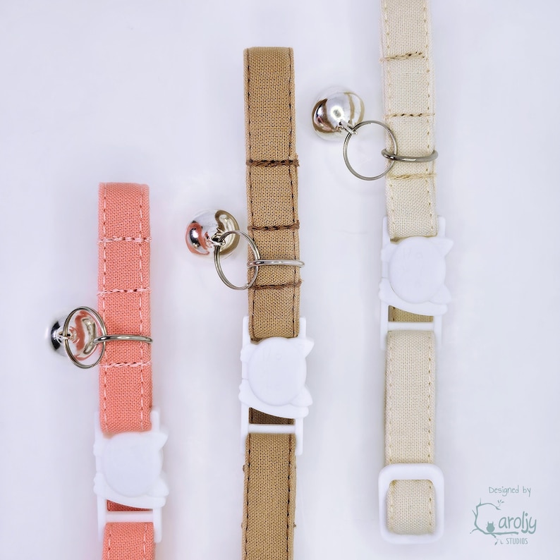 May include: Three fabric pet collars in coral, tan, and cream colors. Each collar features a silver-toned metal bell attached to a split ring, a white plastic buckle shaped like a cat head, and a white plastic adjustment slider for sizing.