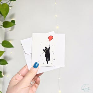 May include: A white greeting card featuring a black cat holding a red balloon, with small black dots scattered around. The card is paired with a white envelope. The design is simple and whimsical.