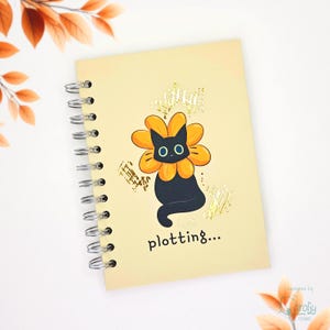 May include: A spiral-bound notebook with a light yellow cover featuring a black cat wearing an orange flower collar. The design includes gold foil accents and the text plotting... printed in black at the bottom of the front cover.