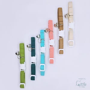 May include: Six fabric pet collars in lime green, dark teal, bright turquoise, coral pink, tan, and cream. Each collar features a white plastic breakaway buckle, a silver metal D-ring, and a small silver bell. The text Caroljy Studios is visible.