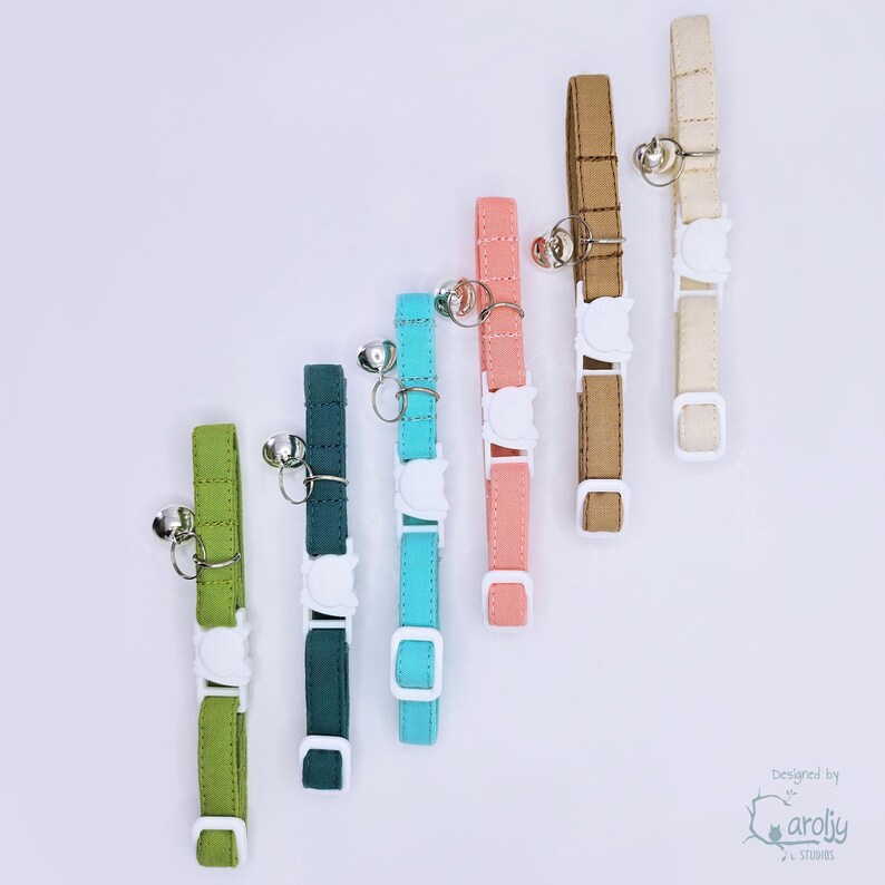 May include: Six fabric pet collars in lime green, dark teal, bright turquoise, coral pink, tan, and cream. Each collar features a white plastic breakaway buckle, a silver metal D-ring, and a small silver bell. The text Caroljy Studios is visible.