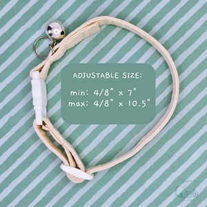 May include: A cream-colored pet collar with a silver-toned metal bell and a white plastic buckle. The text on the image reads: ADJUSTABLE SIZE: min: 4/8" x 7" max: 4/8" x 10.5". The collar features a sliding white plastic adjuster for sizing.