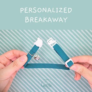 May include: A teal fabric breakaway collar featuring a white plastic cat-shaped buckle, a white plastic adjuster, and a small silver metal bell. The text PERSONALIZED BREAKAWAY is printed in white above the collar against a light blue background.