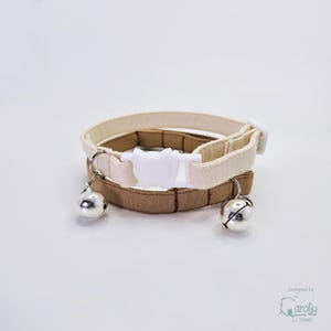 May include: Two pet collars, one in cream and one in tan, featuring a white plastic cat-shaped buckle closure. Each collar is equipped with a small, round, silver-colored metal bell attached to a metal ring, providing a clear audible chime.
