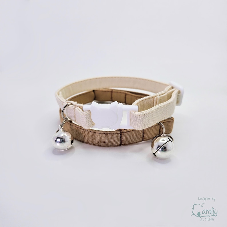 May include: Two pet collars, one in cream and one in tan, featuring a white plastic cat-shaped buckle closure. Each collar is equipped with a small, round, silver-colored metal bell attached to a metal ring, providing a clear audible chime.