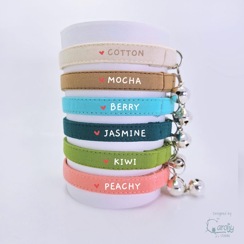 May include: A stack of six fabric collars in cream, mocha, berry, jasmine, kiwi, and peachy colors. Each collar features a small red heart icon, a printed name, and a silver metal bell attached to the side. The names are Cotton, Mocha, Berry, Jasmine, Kiwi, and Peachy.