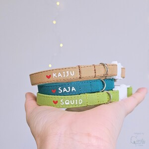 May include: Three fabric pet collars in tan, teal, and light green. Each collar features a small red heart icon, white text reading KAIJU, SAJA, or SQUID, a silver metal D-ring, and a white plastic side-release buckle for secure fastening.