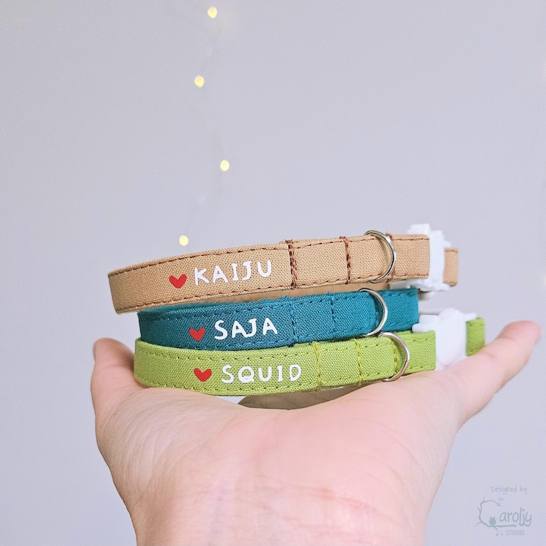 May include: Three fabric pet collars in tan, teal, and light green. Each collar features a small red heart icon, white text reading KAIJU, SAJA, or SQUID, a silver metal D-ring, and a white plastic side-release buckle for secure fastening.