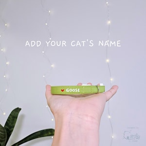 May include: A lime green cat collar featuring the white text GOOSE preceded by a small red heart icon. The collar includes a metallic D-ring for attachment and is presented with the text ADD YOUR CAT'S NAME displayed above the product.
