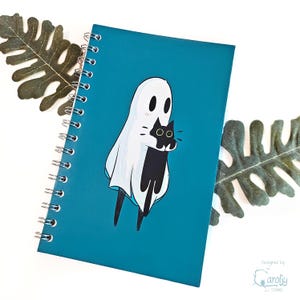 May include: A teal spiral-bound notebook featuring a whimsical illustration of a ghost embracing a black cat. The ghost is white with black eyes, and the cat has large, round eyes. The notebook has a silver spiral binding and the text "designed by Carolij Studios".