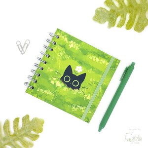 May include: A spiral-bound notebook with a green cover featuring a black cat design and a white flower. A green pen and a silver paperclip are next to the notebook. The notebook has a green elastic band closure.