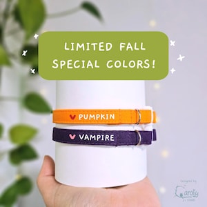 May include: Two fabric collars wrapped around a white cylinder. The top collar is orange with a red heart and the text PUMPKIN. The bottom collar is dark purple with a pink heart and the text VAMPIRE. A green banner above reads LIMITED FALL SPECIAL COLORS!