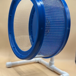 May include: A blue and silver hamster wheel with a mesh interior and a blue plastic rim. The wheel is supported by a white PVC pipe frame, designed for small pets to exercise.