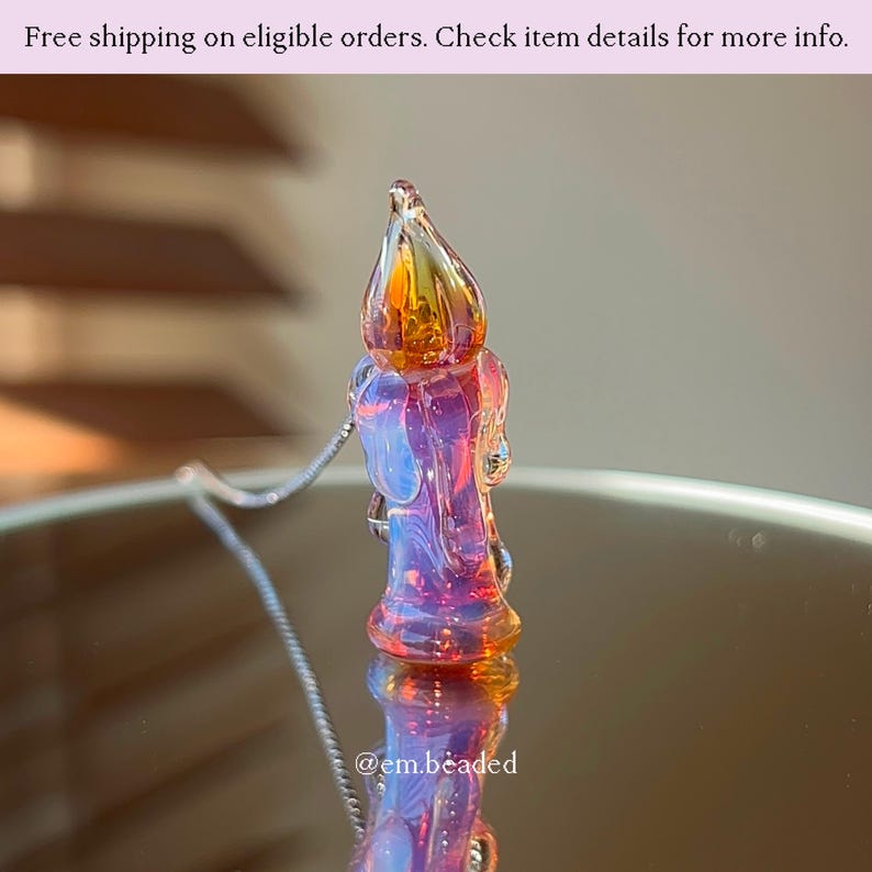 K&ouml;nnte beinhalten: Halskette mit einem Glasanh&auml;nger in Kerzenform. Der Anh&auml;nger hat eine Flamme in Orange und Gelb, mit Rosa- und Blaut&ouml;nen. Die Kette hat eine silberne Kette. Der Text "@em.beaded" ist sichtbar.