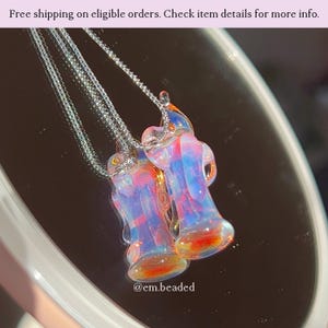 K&ouml;nnte beinhalten: Zwei Glas-Anh&auml;nger-Halsketten mit farbenfrohen, abstrakten Designs in Rosa-, Blau- und Oranget&ouml;nen. Die Anh&auml;nger h&auml;ngen an filigranen Silberketten. Das Bild enth&auml;lt den Text "@em.beaded".
