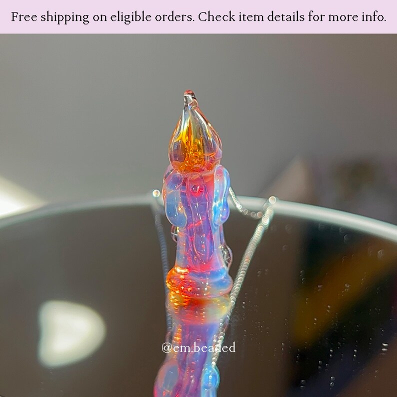 K&ouml;nnte beinhalten: Ein farbenfroher Glasanh&auml;nger in Flammenform mit Orange-, Gelb-, Rot- und Blaut&ouml;nen. Der Text "@em.beaded" ist sichtbar. Der Anh&auml;nger ist wahrscheinlich ein handgefertigtes Schmuckst&uuml;ck.
