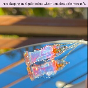 K&ouml;nnte beinhalten: Zwei schillernde Glasanh&auml;nger mit floralem Design, die sich auf einer spiegelnden Oberfl&auml;che spiegeln. Die Anh&auml;nger haben einen transparenten K&ouml;rper mit orangefarbenen und rosa Akzenten. An jedem Anh&auml;nger ist eine silberne Kette befestigt. Der Text "@em.beaded" ist sichtbar.