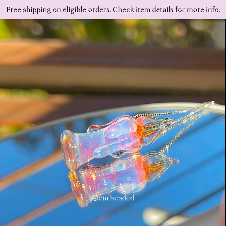 K&ouml;nnte beinhalten: Zwei schillernde Glasanh&auml;nger mit floralem Design, die sich auf einer spiegelnden Oberfl&auml;che spiegeln. Die Anh&auml;nger haben einen transparenten K&ouml;rper mit orangefarbenen und rosa Akzenten. An jedem Anh&auml;nger ist eine silberne Kette befestigt. Der Text "@em.beaded" ist sichtbar.