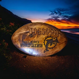 May include: A brown leather football with the words "Miami Dolphins" and a dolphin logo. The football is sitting on a sandy beach at sunset.