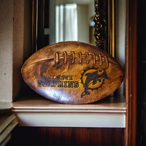 May include: A wooden football with the Miami Dolphins logo and team name. The football is brown and has white stitching.