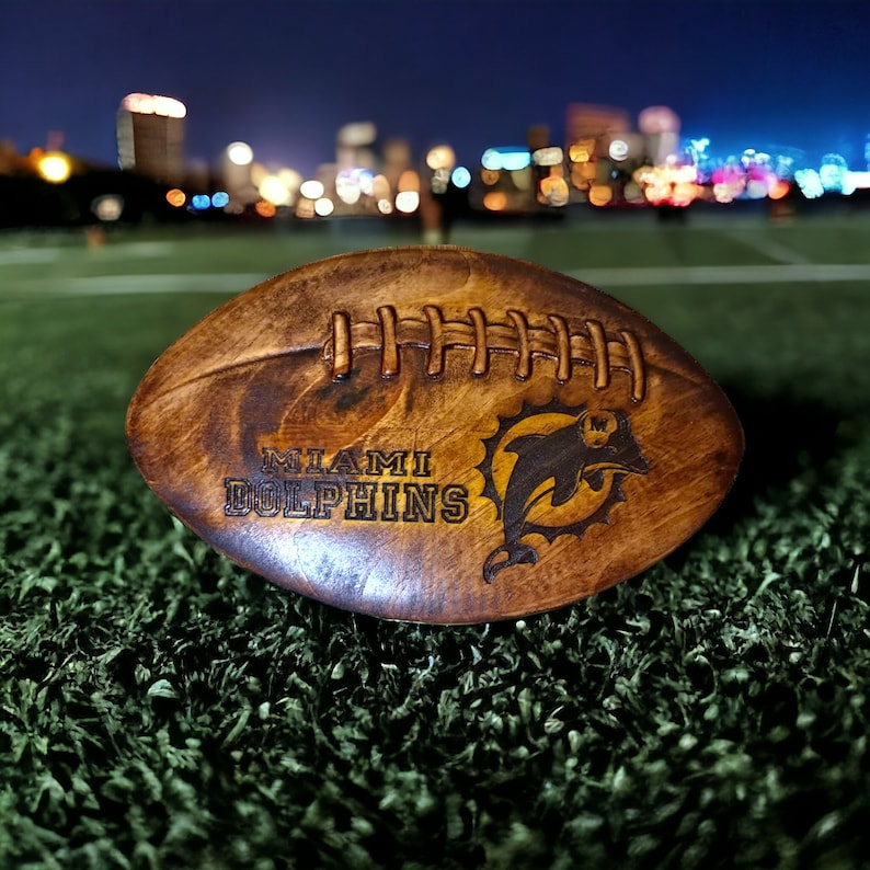 May include: A brown leather football with the words "Miami Dolphins" and a dolphin logo. The football is on a green artificial turf surface.