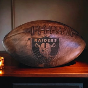May include: A dark brown wooden football with the Raiders logo carved into the side. The logo is a black and white shield with the word "RAIDERS" in white letters and a stylized football helmet with a face mask.