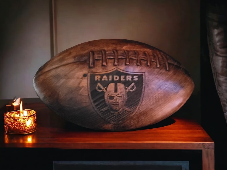 May include: A dark brown wooden football with the Raiders logo carved into the side. The logo is a black and white shield with the word "RAIDERS" in white letters and a stylized football helmet with a face mask.
