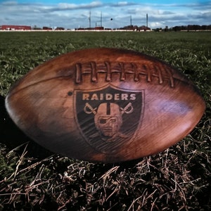 May include: A brown wooden football with the Raiders logo and the word "RAIDERS" in white lettering.
