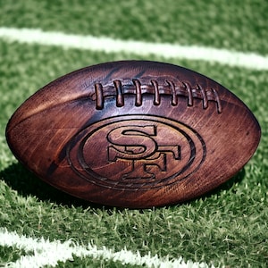 Peut inclure: Un ballon de football américain en bois marron avec le logo des 49ers de San Francisco gravé. Il repose sur un terrain vert avec des lignes blanches. Le logo est un "SF" stylisé dans un ovale rouge.