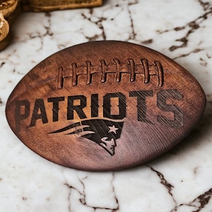Könnte beinhalten: Ein Untersetzer aus Holz in Form eines Fußballs mit dem eingravierten Wort "PATRIOTS". Der Untersetzer zeigt ein stilisiertes Logo der New England Patriots.