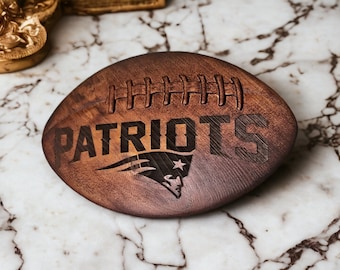 May include: A wooden football-shaped coaster with the word "PATRIOTS" engraved on it. The coaster features a stylized New England Patriots logo.