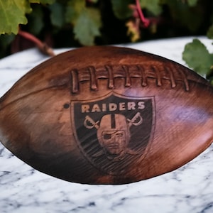 May include: A brown leather football with the Las Vegas Raiders logo engraved on the side. The logo is a black and white shield with the team name "RAIDERS" and a pirate skull with crossed swords.