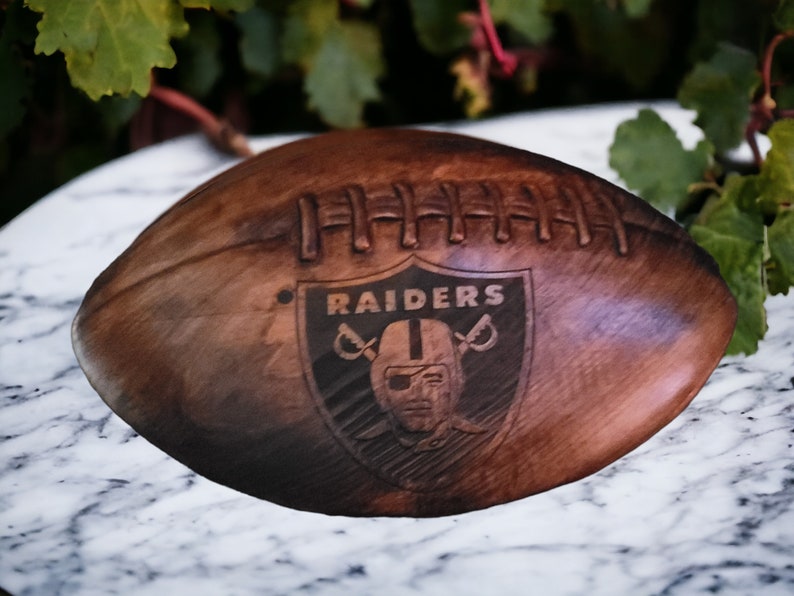 May include: A brown leather football with the Las Vegas Raiders logo engraved on the side. The logo is a black and white shield with the team name "RAIDERS" and a pirate skull with crossed swords.