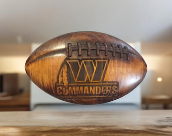 May include: A wooden football with a brown stain finish. The football has a stylized "W" with a feather design and the word "Commanders" engraved on it.