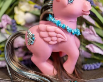 May include: A pink toy pony with blue and brown hair, a turquoise necklace, and a butterfly-shaped emblem on its flank. The pony has wings and is posed on a wooden surface, with a floral background. The logo "Whisper's Vintage Ponies" is visible.