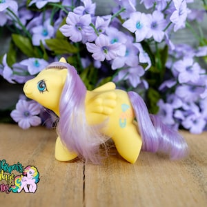 Puede incluir: Un poni de juguete amarillo con pelo y alas moradas, sobre un fondo de flores moradas. El poni tiene ojos azules y un diseño colorido en el flanco. El logotipo "Whisper's Vintage Ponies" es visible en la esquina inferior izquierda.