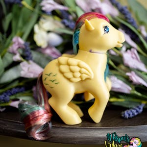 Puede incluir: Un poni de juguete amarillo vintage con melena y cola arcoíris, ojos azules y alas. El poni tiene siluetas de pájaros verdes en el flanco. La imagen incluye un logotipo que dice "Whisper's Vintage Ponies".