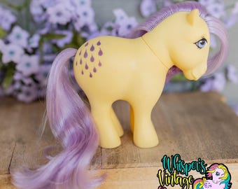 May include: A yellow plastic toy pony with a long, flowing lavender mane and tail. The pony features a purple raindrop pattern on its flank and large blue eyes. The text Whisper's Vintage Ponies is visible in the bottom right corner.